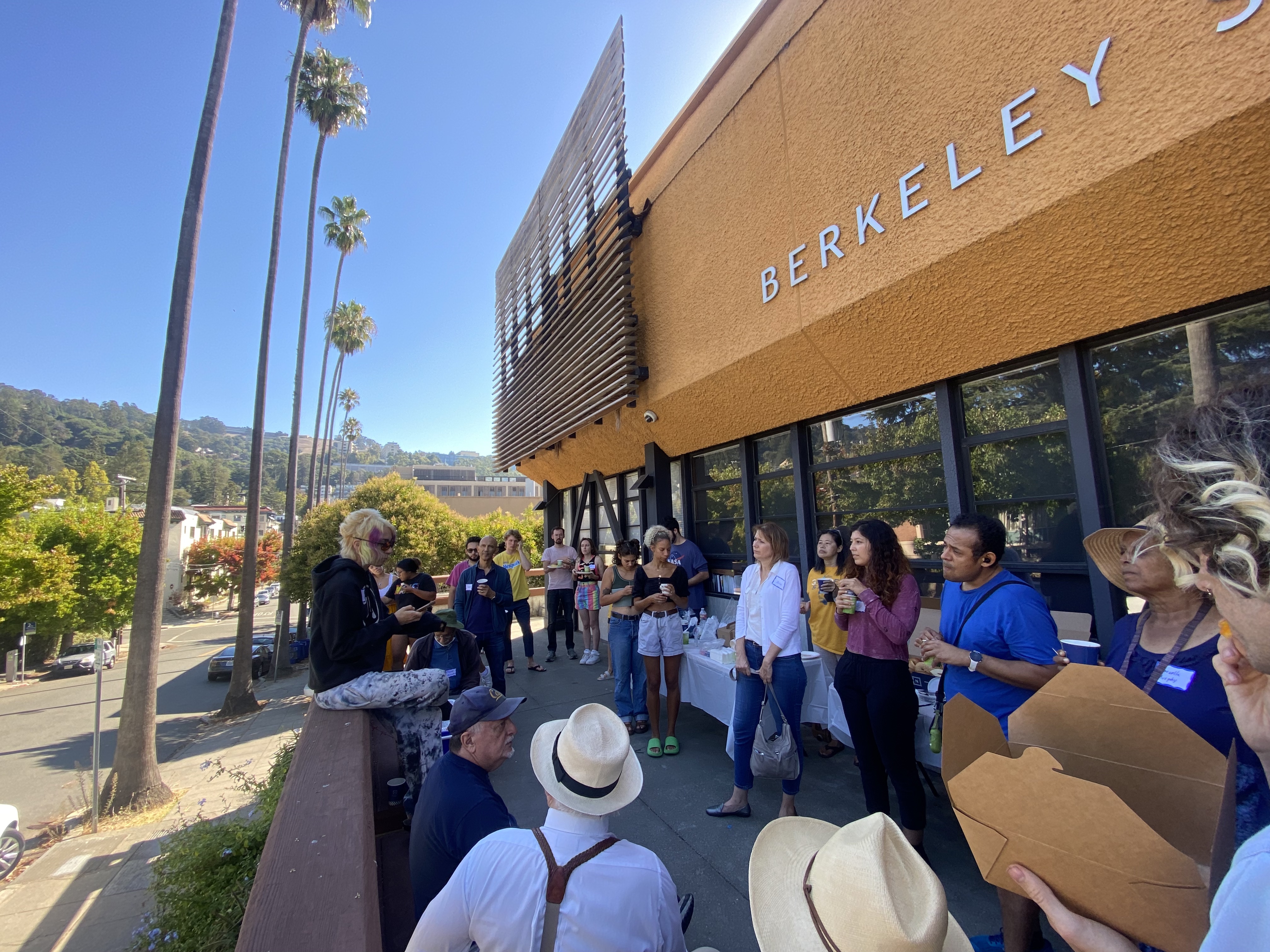 People gathered outside the central office on a clear, sunny day celebrating the 90th anniversary of the Berkeley Student Cooperative with the Berkeley hills in the background